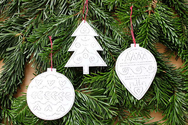 Geprickelte Anhänger in Form von Weihnachtskugel, Weihnachtsbaum und Zapfen hängen an Tannenzweigen